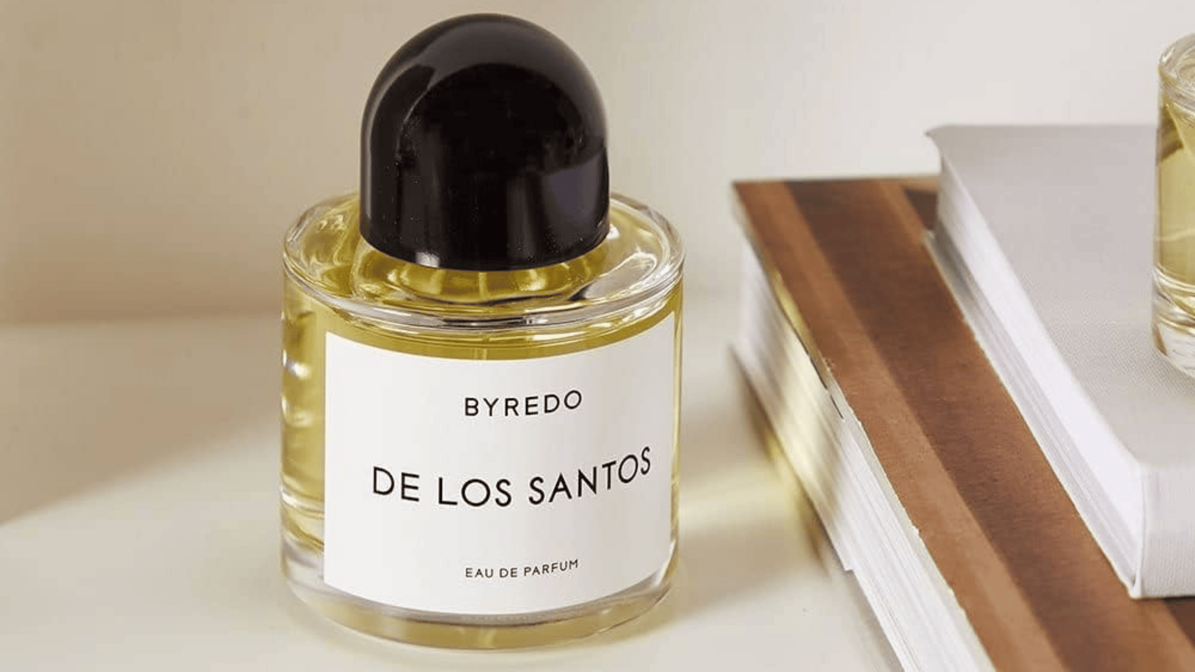 En flaska Byredo De Los Santos Eau de Parfum placerad på en hylla bredvid några böcker. Flaskan har en elegant, minimalistisk design med svart kork och en klar etikett med svart text. Parfymen speglar en känsla av exklusivitet och stilren estetik.